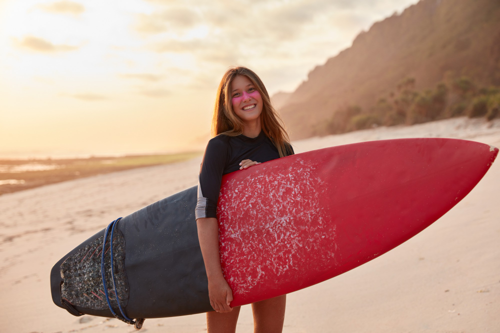 A smiling young woman holding a red surfboard on a scenic sandy beach at sunset, with cliffs and greenery in the background.