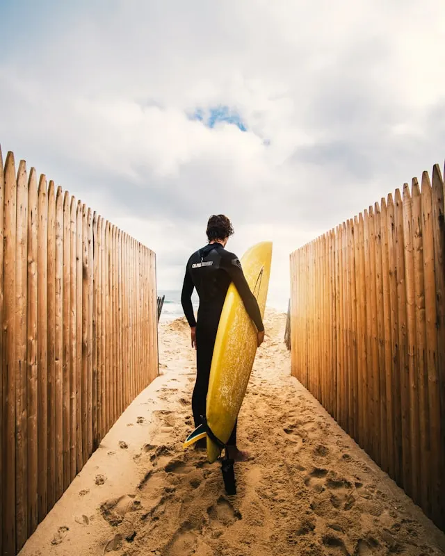 A surfer in a black wetsuit walking down a sandy pathway between wooden fences, carrying a yellow surfboard, heading toward the beach under a cloudy sky.