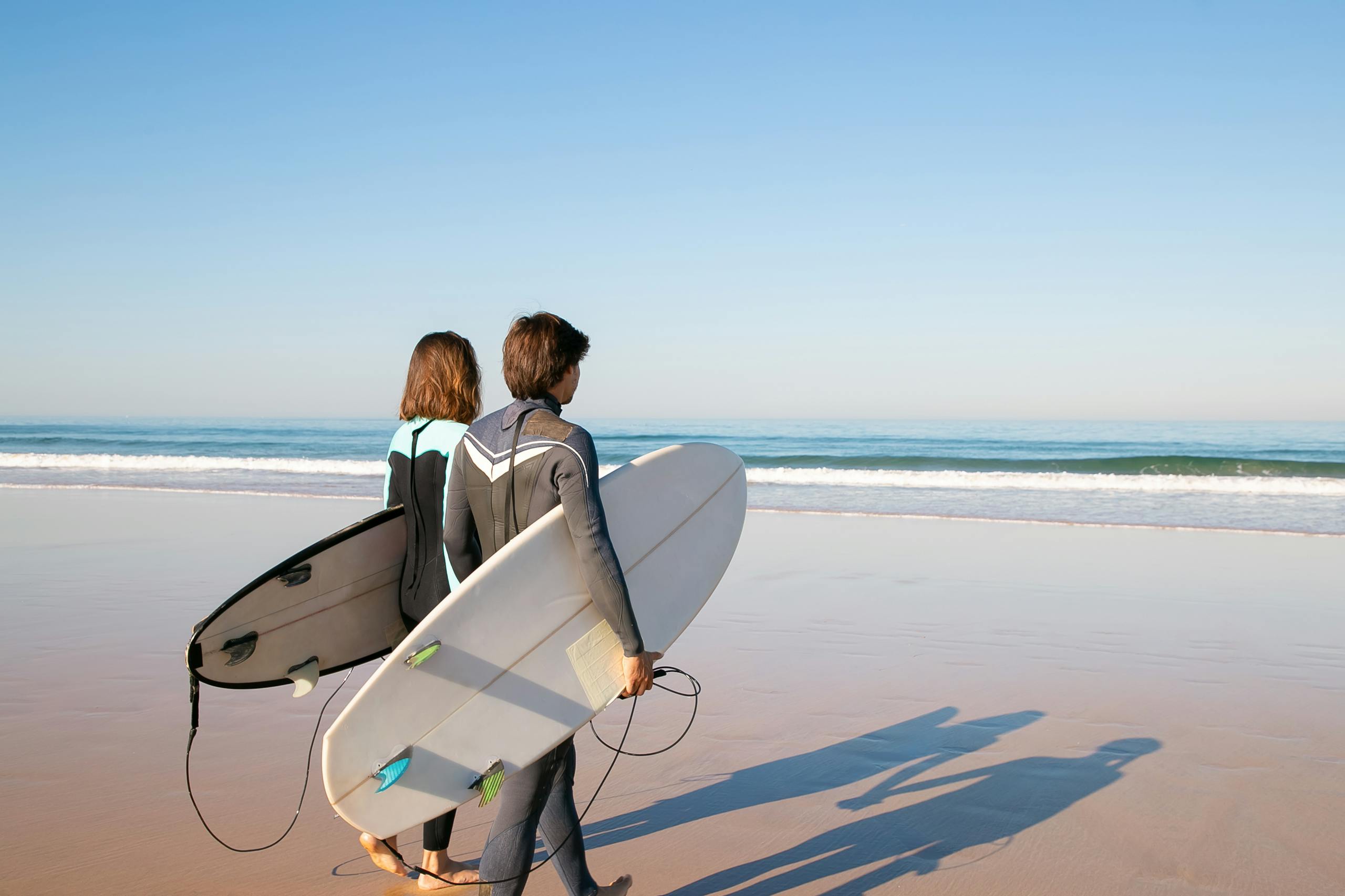 Service 2 A young couple in wetsuits carrying surfboards along a sunny beach in Portugal.